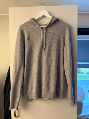 Ljusgrå hoodie i mjukt material - Säljer en stilren ljusgrå hoodie med huva och dragsko. Hoodien har långa ärmar och ribbade muddar vid ärmslut och nederkant. Tillverkad i ett mjukt, stickat material som känns skönt mot huden. Perfekt för en avslappnad look.