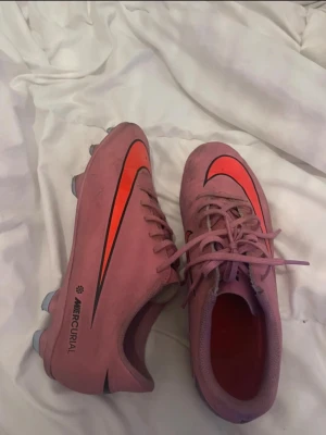 Rosa Nike Mercurial fotbollsskor - Säljer ett par rosa Nike Mercurial fotbollsskor med orange och svart swoosh på sidan. Skorna har klassisk snörning och dobbar för spel på gräs. Snygg och sportig design som sticker ut på planen.