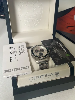 Äkta Certina herrklocka i silver och svart - Snygg Certina herrklocka med robust armband i silverfärgat stål och urtavla i vitt och svart. Klockan har kronograf, tydliga markeringar och sportig design. Perfekt för dig som gillar stilrena accessoarer med funktionella detaljer.