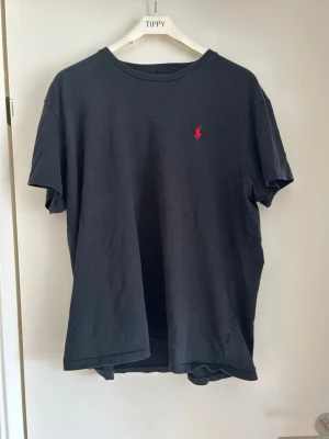 Svart t-shirt med röd logga - En svart t-shirt i klassisk modell med rund hals och korta ärmar. På bröstet finns en liten röd broderad logga som ger en snygg detalj. Perfekt till jeans eller shorts för en enkel och stilren look.   Priset är inte skrivet i sten!!!