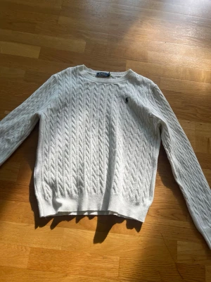 Vit kabelstickad tröja från Ralph Lauren - Kabelstickad vit tröja från Ralph Lauren med klassisk rund halsringning och broderad marinblå logga på bröstet. Tröjan har långa ärmar och ribbade muddar vid ärmslut och nederkant. Perfekt för en stilren och tidlös look.