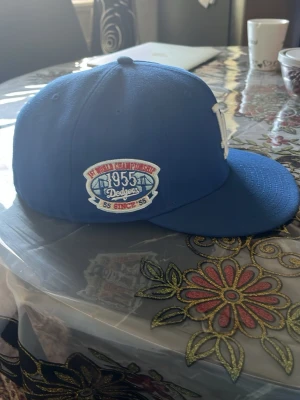 Blå LA Dodgers keps med brodyr - Snygg blå keps med platt skärm och broderad LA-logga framtill. På sidan finns ett märke med texten '1955 Dodgers' och 'World Championship'. Perfekt för dig som gillar sportig stil och klassiska basebollkepsar.