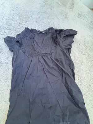 Svart blus med volangkrage XS - Svart blus från 157 i storlek XS. Blusen har korta ärmar, volangdetaljer vid kragen och ett dekorativt veckat parti framtill. Tillverkad i ett tunt, lätt tyg som ger en luftig känsla. Perfekt för dig som gillar en söt och enkel stil.