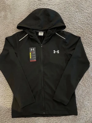 Svart hoodie från Under Armour - Svart hoodie med dragkedja från Under Armour. Stort logotryck på ryggen och mindre logga på bröstet. Huva och långa ärmar, tillverkad i ett sportigt funktionsmaterial. Perfekt för träning eller chill.