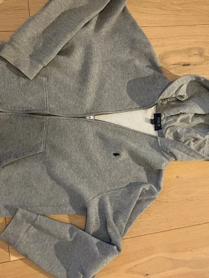 Grå zip hoodie från Polo Ralph Lauren - Säljer en grå hoodie med dragkedja från Polo Ralph Lauren. Klassisk design med huva, två fickor framtill och den ikoniska lilla loggan broderad på bröstet. Tillverkad i mjukt sweatshirtmaterial som är perfekt för chill dagar. Lite trög drag kedja 