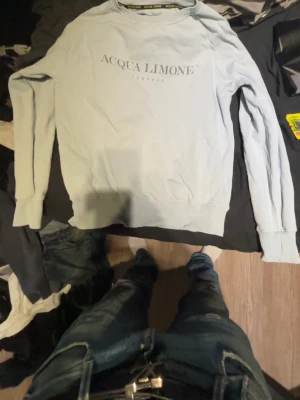 Ljusblå sweatshirt från Acqua Limone - Ljusblå sweatshirt från Acqua Limone med tryck på bröstet och en stor gul citronlogga på ryggen. Tröjan har rund halsringning, långa ärmar och breda muddar vid ärmslut och nederkant. Perfekt för en avslappnad och trendig look.