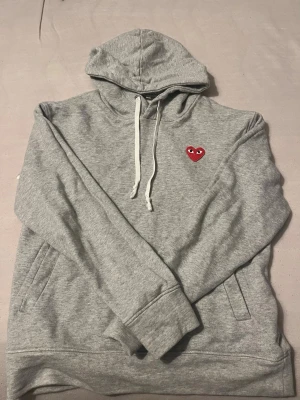 Grå cdg hoodie  - Snygg grå hoodie med huva och vita dragsnören. På bröstet finns ett broderat rött hjärta med ögon, vilket ger en unik och trendig look. Hoodien har känguruficka och är tillverkad i mjukt material, perfekt för en avslappnad stil.