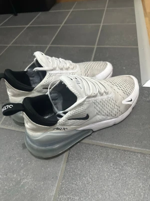 Vita Nike Air Max 270 sneakers - Snygga vita Nike Air Max 270 sneakers med svart swoosh och detaljer. Ovandelen är i mesh för bra ventilation och sulan har en tydlig Air-enhet i hälen för extra komfort. Skorna har svarta insidor och dragflik bak med texten 'AIR270'. Perfekta för en sportig look.