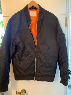 Svart bomberjacka från Jack & Jones - Svart bomberjacka från Jack & Jones med orange foder och klassisk silhuett. Jackan har dragkedja framtill, två snedställda fickor med tryckknapp och ribbade muddar vid ärmslut och midja. Perfekt för en cool streetstil.