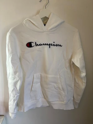 Vit hoodie från Champion med logga - Säljer en vit hoodie från Champion med stor broderad logga i svart, rött och vitt på bröstet. Klassisk känguruficka framtill och ribbade muddar. Skulle säga att det är en storlek xs. Är en gul fläck på vänster ärm (se sista bilden), men ingenting som förstör helhetsintrycket. 