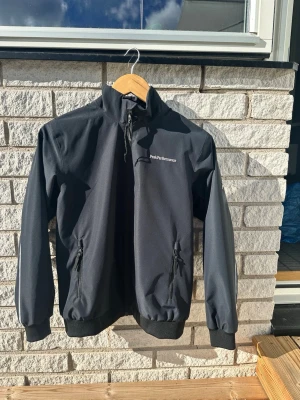 Svart vindjacka från Peak Performance - Snygg svart vindjacka från Peak Performance med dragkedja framtill, två fickor med dragkedja och hög krage med justerbar dragsko. Jackan har ribbade muddar vid ärmslut och nederkant. Perfekt för blåsiga dagar och enkel att matcha. Det är storlek 160
