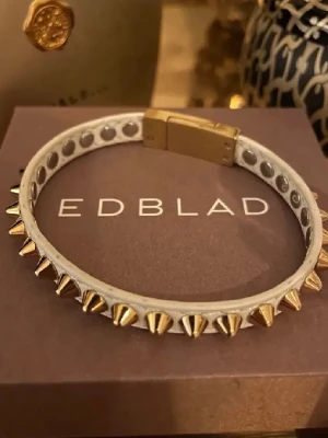 Vitt armband med guldnitar från Edblad - Snyggt vitt nitarmband från Edblad, jättebra skick! Köpt för 399kr o säljer för 199kr💗Armbandet har ett magnetlås i guld som matchar nitarna. Längden är den minsta 16cm