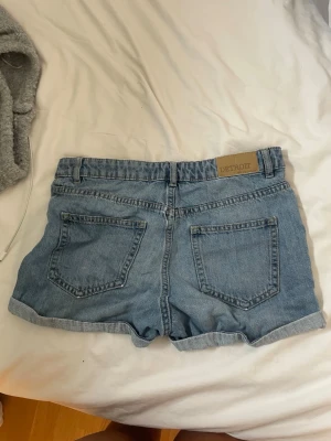 Blå denimshorts från Lindex - Säljer ett par klassiska blå denimshorts från Lindex med uppvikta benslut och femficksmodell. Shortsen har bälteshällor, knapp och dragkedja framtill samt en patch med texten 'DETROIT' bak. Perfekta för sommaren!