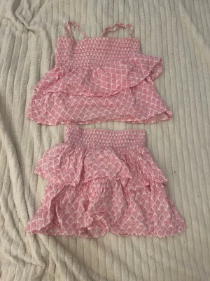 Rosa volangset från Lindex - Sött tvådelat set från Lindex i storlek 158/164. Setet består av ett linne med smala axelband och volang samt en matchande kjol med dubbla volanger. Mönstret är rosa och vitt med små blommor. Perfekt för dig som gillar en romantisk och somrig stil.