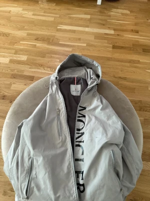 Ljusgrå vindjacka från Moncler - Snygg ljusgrå vindjacka från Moncler med huva och dragkedja framtill. Jackan har stora Moncler-trycket på bröstet, två sidofickor med dragkedja och Moncler-logga på ärmen. Tillverkad i lätt och tåligt material, perfekt för blåsiga dagar.