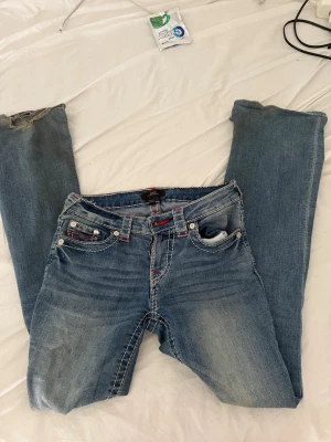 True Religion jeans i modellen 'Becca', storlek S, begagnat skick med synligt slitage vid vänster benslut. - True Religion Becca jeans stl S | Y2K vintage blå röd sömnad True Religion Becca-jeans i storlek S 🙌 Mellanblå tvätt med signaturröd kontrastsömnad — klassisk Y2K-look Fits slim, superbra med ett oversized linne eller croppad hoodie Begagnat skick — visst slitage vid vänster benslut, annars inga synliga defekter Perfekt för dig som bygger upp det där tidiga 2000-tals-outfittet