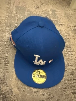 Blå LA keps med brodyr - Snygg blå keps med LA-logga framtill och broderad text 'LOVE & HUSTLE = GAME' på sidan. Kepsen har platt skärm och klassisk 59FIFTY-stil. Materialet är bomull och kepsen har MLB-logga på sidan.
