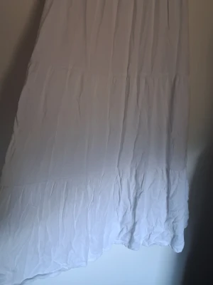 Vit långkjol i bomull - Säljer en flowy vit långkjol med volangdetaljer. Kjolen är sydd i flera lager och har en avslappnad, luftig känsla som passar perfekt till varma dagar. Materialet känns mjukt och lätt mot huden.