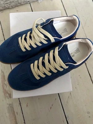 Blå Maison Margiela sneakers - Snygga blå sneakers från Maison Margiela med beige snörning och gummisula. Ovandelen är i mocka och läder, vilket ger en schysst kontrast. Låg modell med vit insida och klassisk siluett. Perfekta för dig som vill ha något stilrent men ändå unikt. Det finns ingen box