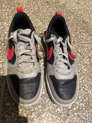 Nike sneakers grå/svart med röd swoosh - Snygga Nike sneakers i lågt modell med grå ovandel, svarta partier och en röd swoosh på sidan. Skorna har gråa snören, svart innersula och vit sula. Perfekta för dig som vill ha en sportig och stilren look. Snygga till vardag med liten pop av färg, säljer på grund av ingen användning, då jag använt de max 5 gånger💕Kostar 650kr ordinarie