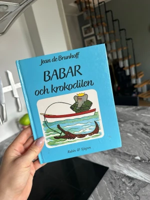 Babar och krokodilen bok - En charmig barnbok med blått hårt omslag, skriven av Jean de Brunhoff. Framsidan visar Babar i en båt som fiskar efter en krokodil. Boken är utgiven av Rabén & Sjögren och ingår i Klumpe Dumpe-serien. Perfekt för dig som gillar klassiska berättelser och fina illustrationer.