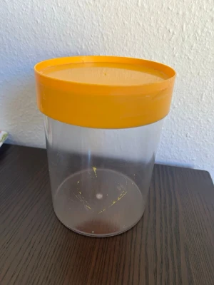 Plastburk med orange lock - En plastburk med ett orange lock. Burken är genomskinlig.  Skick kan ses på bilder!!