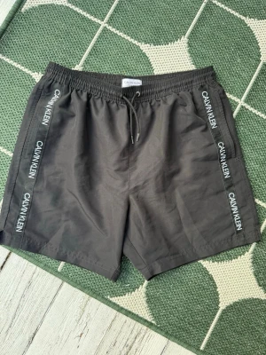 Svarta badbyxor från Calvin Klein - Snygga svarta badbyxor från Calvin Klein med vita loggor längs sidorna och justerbar dragsko i midjan. Tillverkade i snabbtorkande material och har vit mesh på insidan för extra komfort. Perfekta för stranden eller poolen. Fickor fram. Tyvärr har gummibandet i nätinnerbyxan åldrats och har ingen elastisitet kvar, se bild.  Går att klippa bort innerbyxan och köra kalsonger under om man vill. Det står storlek 14-16 år i dessa, så passar barn/ungdom.