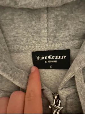Grå Juicy Couture zip hoodie - Mysig grå hoodie från Juicy Couture med dragkedja, huva och dragsnören. Klassisk logga broderad på bröstet och fickor framtill. Perfekt för en chill och avslappnad stil. Tillverkad i mjukt material som känns skönt mot huden.
