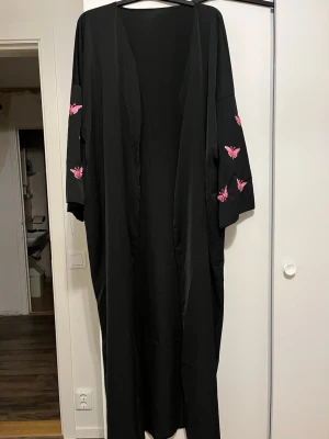 Svart abaya - Lång svart kimono-inspirerad morgonrock med vida ärmar och rosa fjärilar broderade på ärmarna. Morgonrocken har en lös passform och är tillverkad i ett mjukt, lätt material som känns skönt mot huden. Perfekt för att chilla hemma i stil.