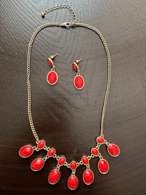 Halsband+ örhängen med röda ovala stenar - Säljer ett halsband+örhängen med kedja i metallfärg och flera ovala röda stenar i olika storlekar. Stenarna är infattade i metall och ger ett statement-utseende. Perfekt för dig som gillar färgstarka accessoarer och vill ha något som verkligen syns.