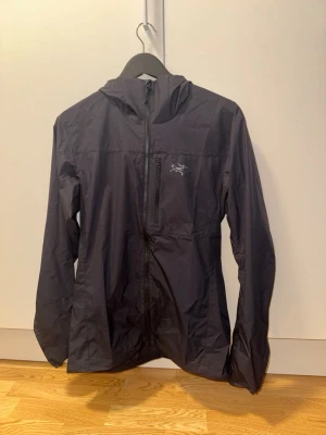 Svart vindjacka med huva Arc'teryx - Snygg svart vindjacka från Arc'teryx med huva och dragkedja framtill. Jackan har en bröstficka med dragkedja och diskret logga på bröstet. Tillverkad i lätt och tunt material som passar perfekt för blåsiga dagar.