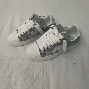 Vita och grå sneakers med camomönster - Snygga sneakers i vitt och grått med camoinspirerat mönster på sidorna. Skorna har breda vita skosnören, vit sula och rund tå. (Pris kan diskuteras😊)