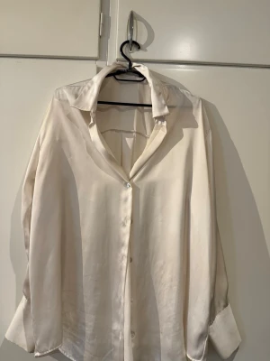 Klassisk vit skjorta från Zara - Stilren vit skjorta från Zara i blankt, mjukt material som ger en lyxig känsla. Skjortan har klassisk krage, långa ärmar med breda manschetter och knäppning framtill. Perfekt för en clean och enkel look.