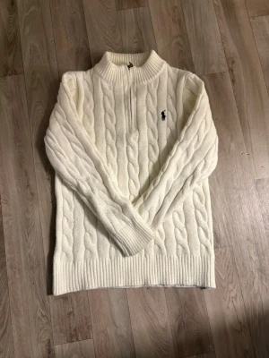 Vit kabelstickad tröja med halv dragkedja - Kabelstickad vit tröja med halv dragkedja och ribbade muddar. Tröjan har en hög krage och ett broderat logotypmärke på bröstet. Perfekt för dig som gillar klassisk och stilren design. Materialet ser ut att vara mjukt och varmt.
