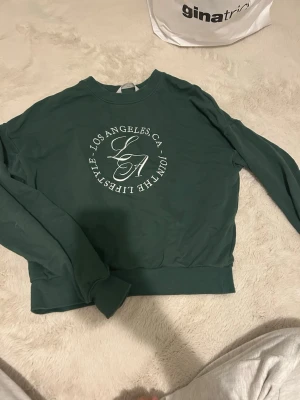 Grön sweatshirt med LA-tryck - Mörkgrön sweatshirt med vitt tryck på bröstet där det står 'Los Angeles CA - Join the Lifestyle'. Tröjan har rund halsringning, långa ärmar och breda muddar vid ärmslut och nederkant. Perfekt för en avslappnad och trendig look.