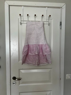 Rosa volangklänning från Gina Tricot - Söt kort klänning från Gina Tricot i ljusrosa och vitt med smock upptill och dubbla volanger nedtill. Klänningen har ett diskret mönster och är perfekt för sommarens varma dagar. Tillverkad i mjuk bomull för en bekväm känsla.