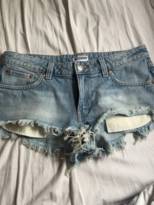 Blå jeansshorts från Weekday - Säljer ett par ljusblå jeansshorts från Weekday med råa, slitna kanter och fransar. Klassisk femficksmodell med knapp och dragkedja framtill. Perfekta för sommaren och ger en avslappnad vibe.