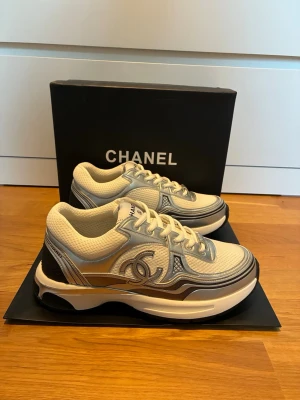 C h a n e l sneakers - Snygga sneakers från C h a n e l i vitt och silver med ikoniska CC-loggan på sidan. Skorna har en chunky sula, mesh- och läderdetaljer samt klassisk snörning. Perfekta för dig som vill ha en exklusiv och trendig look.