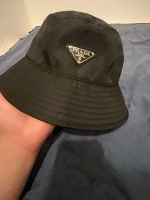 Prada bucket hat - Snygg svart bucket hat från Prada med det klassiska trekantiga metallogot på framsidan. Hatten är tillverkad i ett glansigt, slitstarkt material som ger en exklusiv känsla. Perfekt accessoar för att lyfta din outfit och ge en trendig touch. I gott skick