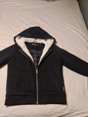 Svart bunny jacka från moose knuckles  - Mysig svart hoodiejacka med vit fuskpäls i huvan och quiltat innerfoder. Jackan har dragkedja, två stora fickor och en avslappnad passform. Perfekt för kyliga dagar när du vill vara både varm och stilren.