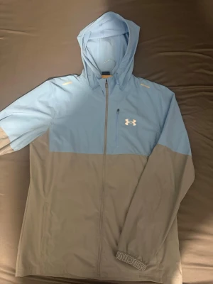 Under Armour tvåfärgad vindjacka - Snygg vindjacka från Under Armour i ljusblått och grått. Jackan har huva, hel dragkedja och en liten bröstficka med dragkedja. Perfekt för blåsiga dagar och sportiga outfits. Logga på bröstet och diskret tryck vid ärmslutet.