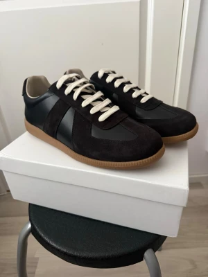 Maison Marigela svarta och bruna 43 - Stilrena svarta sneakers med kontrasterande vita skosnören och beige yttersula. Ovandelen är i en mix av mocka och slätt läder, vilket ger en snygg och modern look. Låg modell som passar perfekt till jeans eller streetwear.