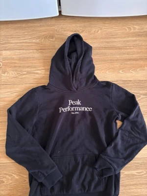 Svart hoodie från Peak Performance - Säljer en svart hoodie från Peak Performance med klassisk logga broderad i vitt på bröstet. Hoodien har stor huva, magficka och ribbade muddar vid ärmar och nederkant. Perfekt för chill dagar eller till skolan. Har en slitning på höger armbåge fast syns inte jätte mycket.