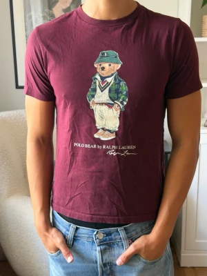 Vinröd Polo Bear t-shirt från Ralph Lauren - Säljer en vinröd t-shirt från Ralph Lauren med den ikoniska Polo Bear-trycket på bröstet. T-shirten har rund hals, korta ärmar och är gjord i mjuk bomull. Perfekt för dig som vill ha en klassisk men lekfull look. 