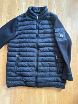 Moncler jacka  - Snygg svart dunjacka från Moncler med quiltad kropp och släta ärmar. Jackan har två dragkedjeförsedda fickor fram och en Moncler-logga på ärmen. Hög krage och rak passform ger en clean look. Perfekt för kyliga dagar. Storlek XL/XXL. 