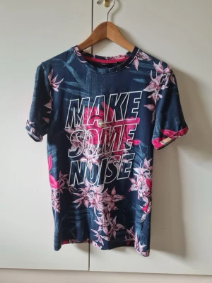 Blommig t-shirt med tryck från CROPP strl S - Snygg t-shirt med stort 'MAKE SOME NOISE'-tryck på bröstet och blommigt mönster i rosa och vitt mot en mörkblå bakgrund. T-shirten har korta ärmar och rund hals. Slim fit. Material 100% bomull. Storlek S.