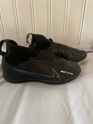 Nike Air Zoom svarta fotbollsskor - Säljer ett par svarta Nike Air Zoom inomhus fotbollskor med låg profil och elastisk strumpa runt ankeln. Skorna har en stilren design med vita och grå detaljer, kardborreband över vristen och slitstark sula för bra grepp på planen.