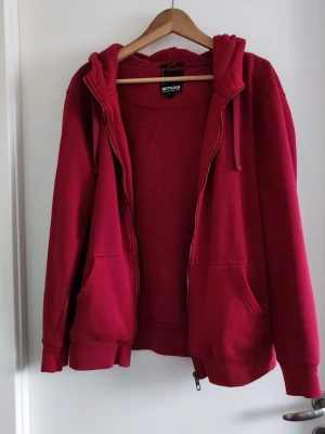 Röd zip hoodie från SMOG - Röd hoodie från SMOG i storlek L med dragkedja, huva med dragsko och två stora fickor framtill. Slim fit-modell i mjukt material, perfekt för chill dagar. Klassisk streetwear-look med ribbade muddar vid ärmar och nederkant.