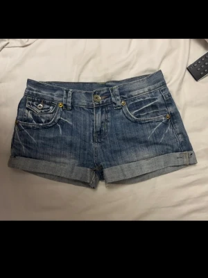 Blå denimshorts med uppvik - Säljer ett par klassiska blå denimshorts med uppvikta benslut och slitna detaljer. Shortsen har fem fickor, bälteshällor och knappgylf. Perfekta för varma dagar och en avslappnad stil. Mått visas på bild. Står ingen storlek men skulle säga xs/s
