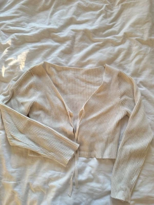 Beige ribbad kofta med knytband - Trendig beige ribbad kofta med långa ärmar och knytband framtill. Croppad modell med djup v-ringning och mjukt, stretchigt material. Perfekt att styla över ett linne eller topp för en avslappnad look.